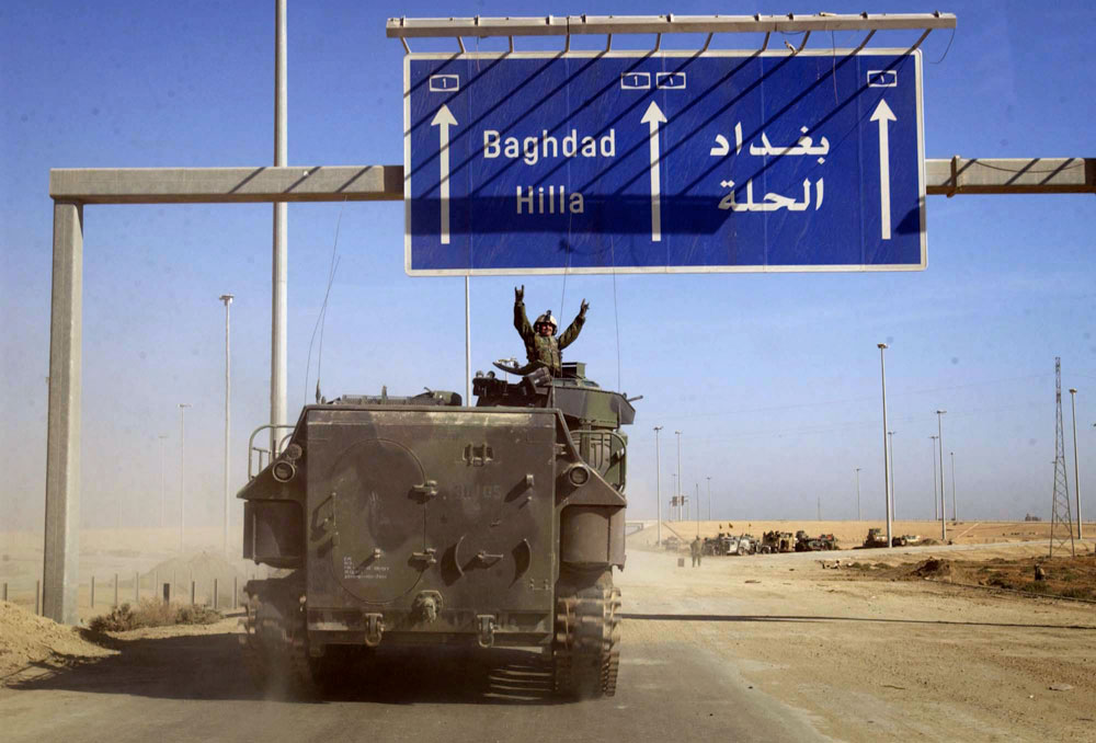 A U.S. Marine raises his arms in triumph as he passes under the first sign for Baghdad on the road north from Kuwait.