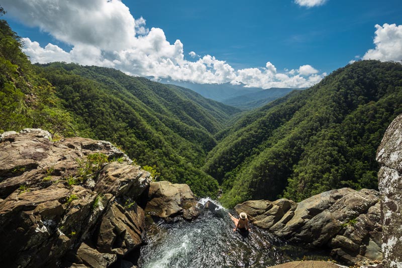 Woman relaxing in a natural pool at the top of a waterfall with views over valley below  