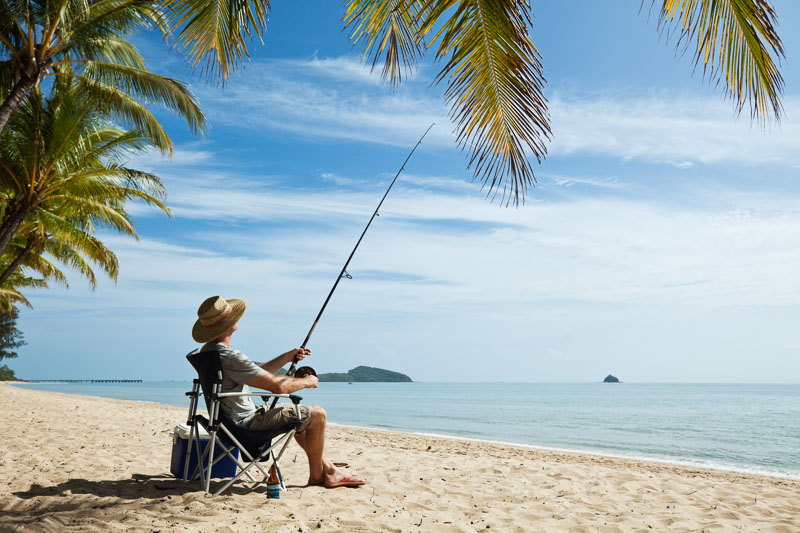 Man sitting in a chair at tropical beach holding a fishing rod 
