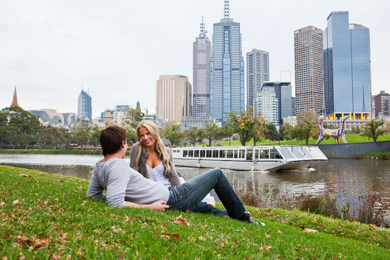 Young couple relaxing on the grassy riverside with city skyline beyond 