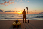 A man with paddle standing next to a kayak looking out to sea at sunrise