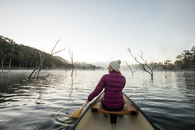 View of a woman paddling a canoe on a misty lake at sunrise  
