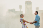 bride and groom smile and hold hands with mist from Niagara Falls and downtown buildings behind them at sunset during their wedding engagement photography session 