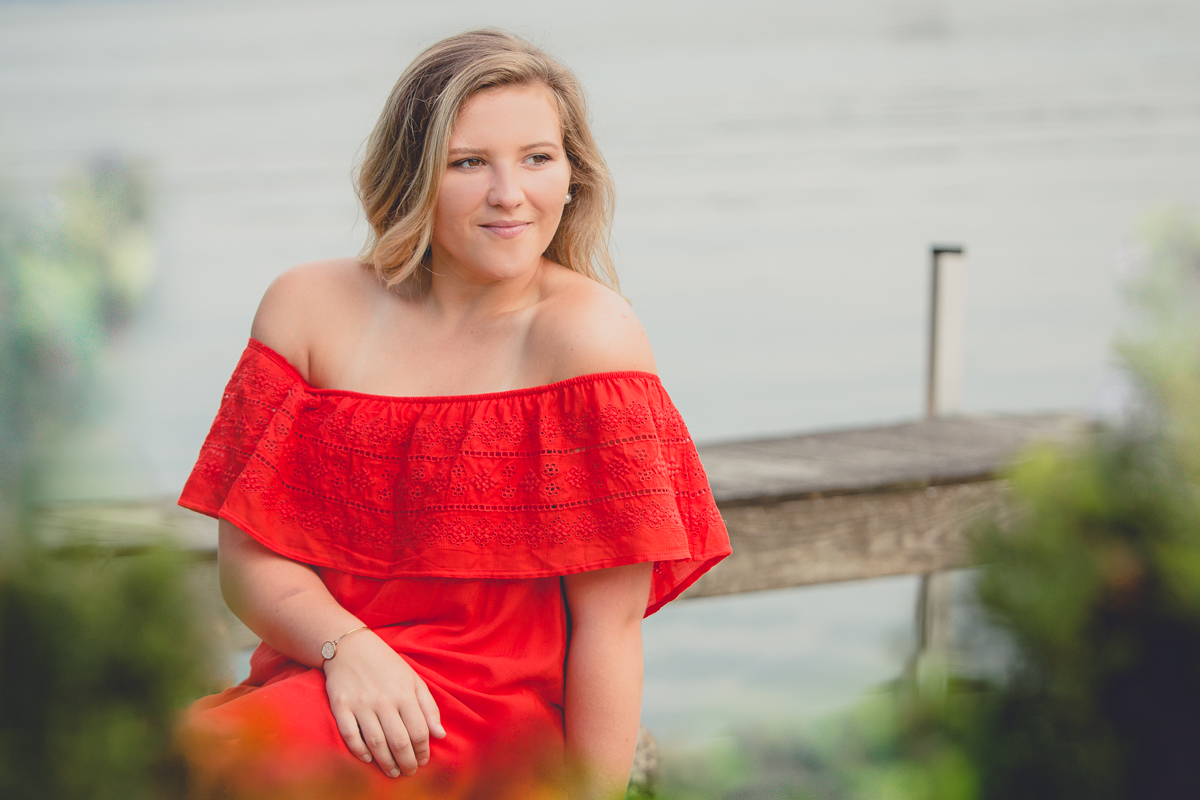 senior portrait by photographer Lindsay DeDario of maple grove high school student sitting in red dress by dock overlooking Lake Chautaqua in Bemus Point, a small town near Buffalo, NY in WNY 
