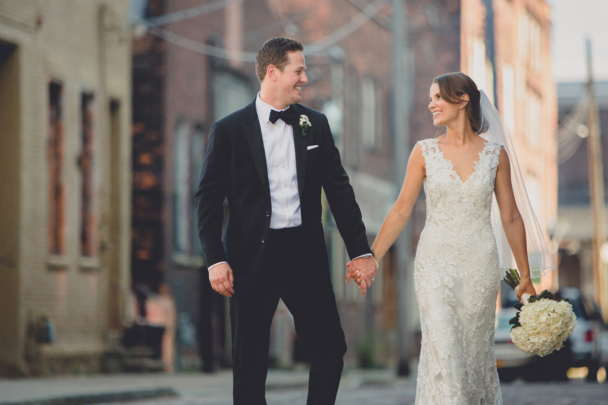 bride and groom smile and walk holding hands down cobblestone street in downtown Buffalo, NY on their wedding day