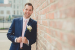 groom leans on brick wall and smiles for photographer on wedding day next to Greetings from Buffalo Mural in Buffalo, NY