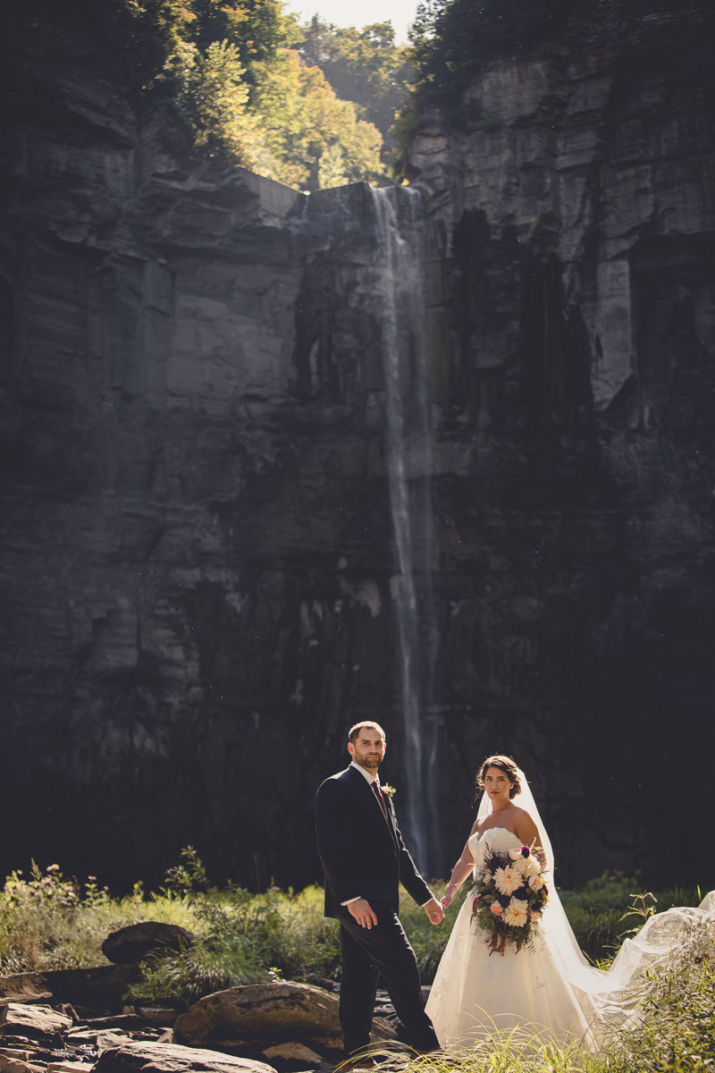 bride and groom stand holding hands and looking at camera in front of Taughannock Falls after their Cornell Sage Chapel wedding ceremony and before heading to Muranda Cheese barn for their reception in Ithaca, NY