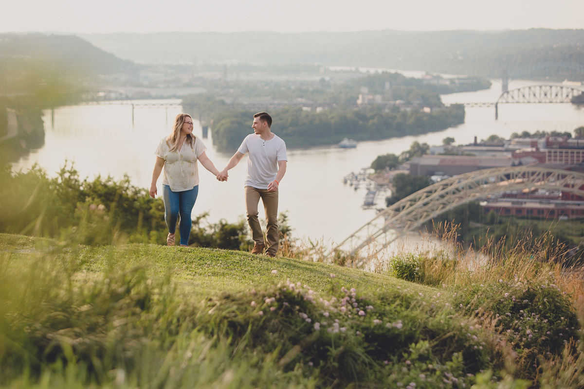 couple holds hands near incline rail in grass overlooking rivers and bridges in Pittsburgh, PA during their engagement photography portrait session