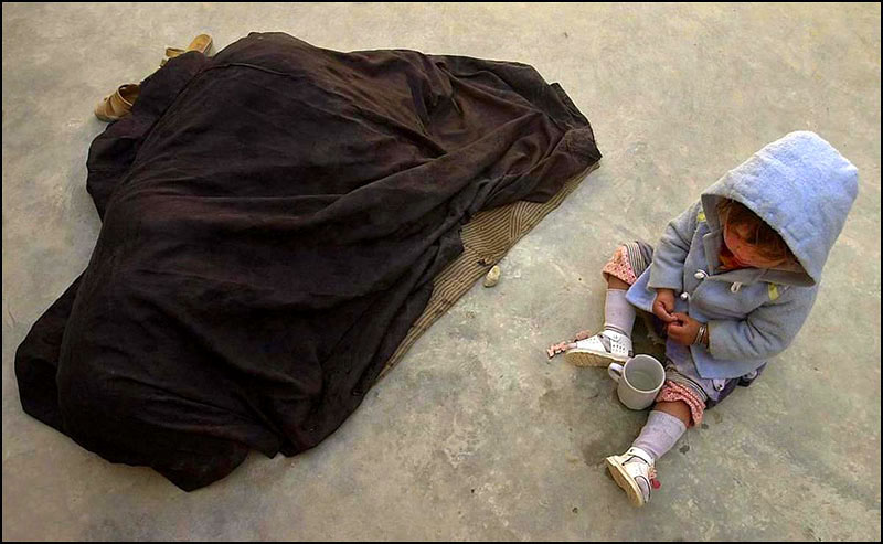 A mother lies covered under a black cloth shaking with sickness as her three-year-old daughter plays with a cup.