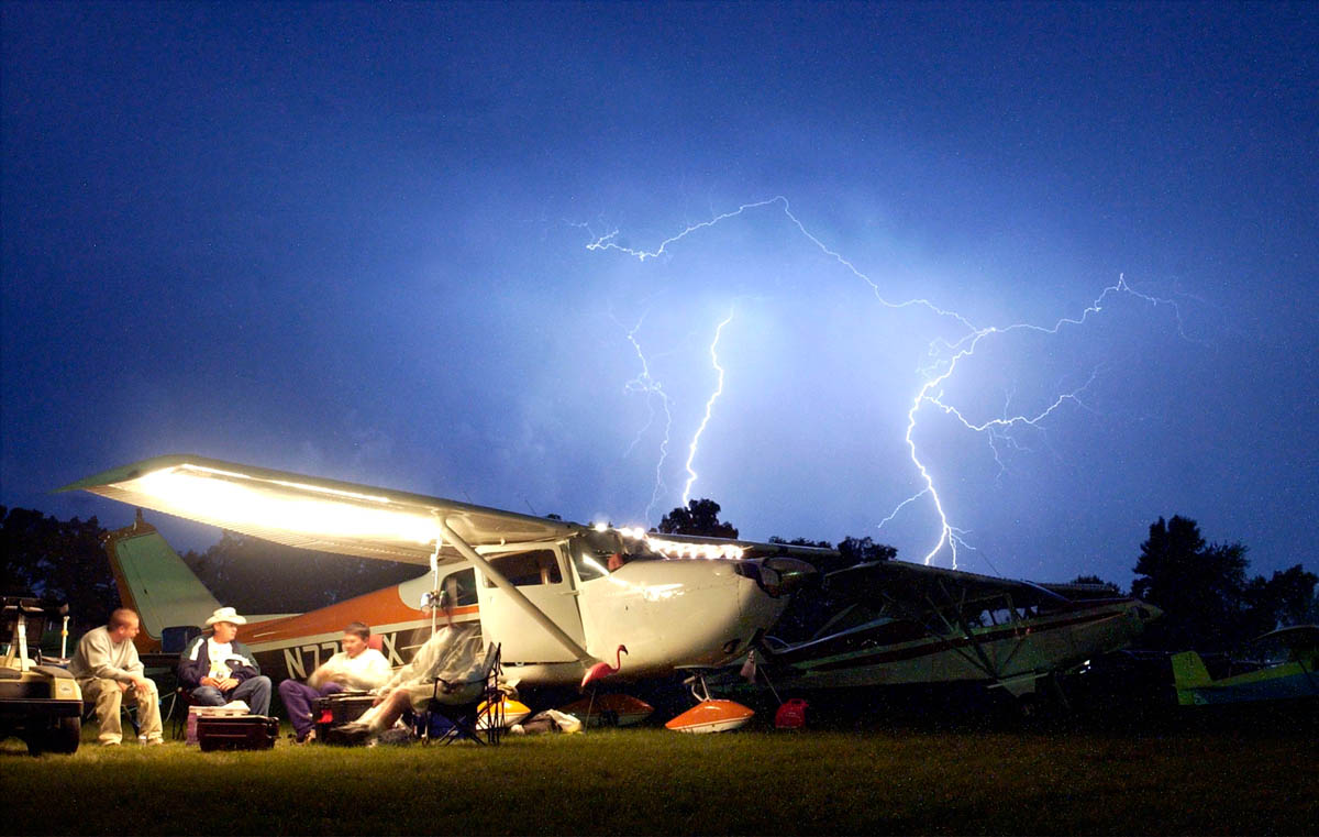 Friends stay dry under the wing of Dan Nelson's 1961 Cessna 172 as lightning strikes and the storms hit the EAA AirVenture in Oshkosh, Wis.