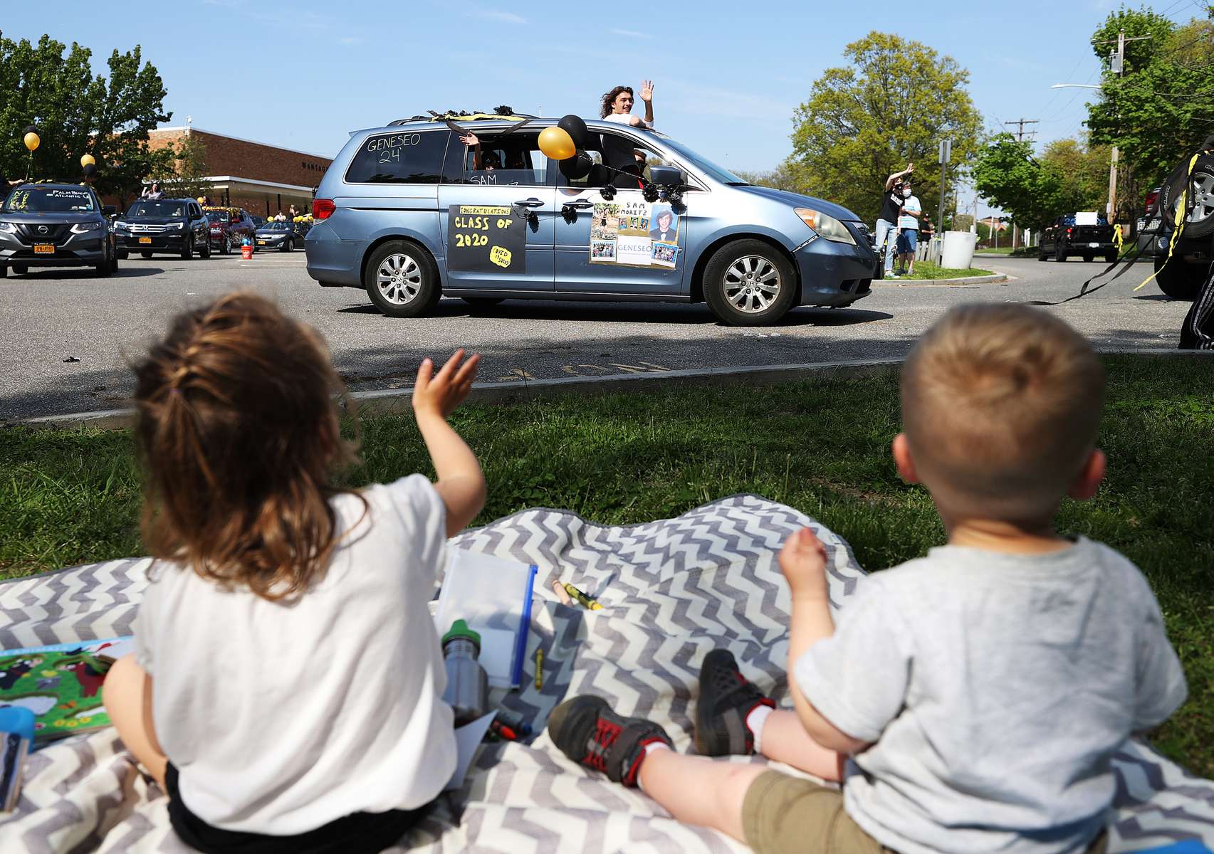 Children wave as graduating Seniors from Wantagh High School drive by the front of the school as their teachers cheer for them on May 15, 2020 in Wantagh, New York.  The school semester  has been cancelled and students had to finish their year with online video classes due to the coronavirus COVID-19 pandemic.  