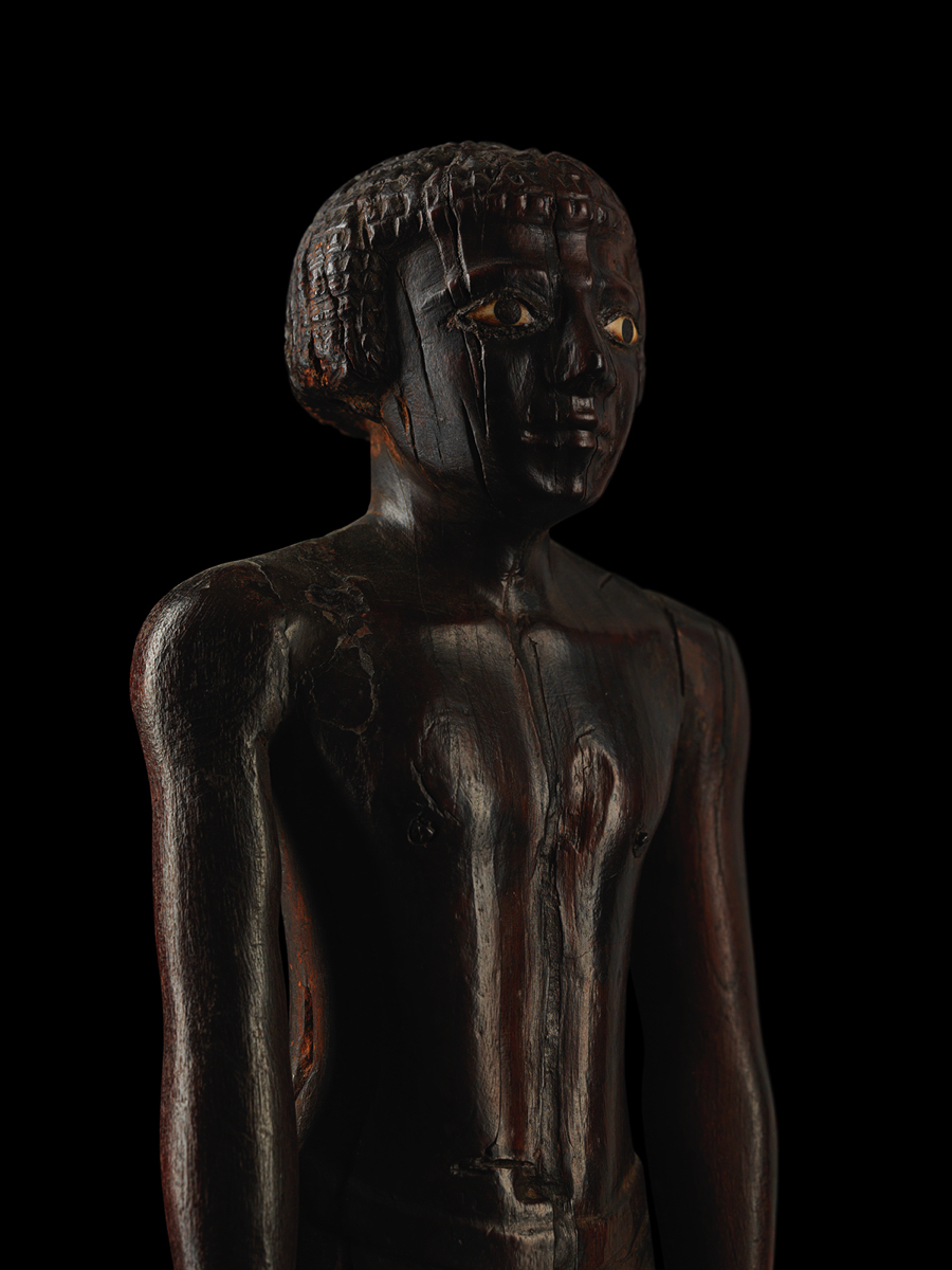 MIDDLE KINGDOM EGYPT (2050-1710 B.C.)Acacia wood, bronze, boneH: 35.8cm This exceptional wooden statue of a man is an exquisite example of the skill with which Egyptian sculptors executed their works. It is constructed from different elements, with the face and both arms being worked separately from the body. In the forehead a dowel is visible, used to connect the facial mask to the rest of the head. Originally, the figure would have been covered in a very thin layer of plaster stucco and brightly painted.The slender figure is shown in a strong vertical pose, with his back straight and head held high. The short round wig the man is wearing, leaves his earlobes visible. The most striking feature of the delicately carved face are the carefully detailed eyes. Within a contour of bronze, the original bone inlay is preserved. Round pupils have been added in black paint. Both arms are held alongside the torso, the hands balled in fists. The right hand turns slightly outwards and is perforated to hold an attribute which is now missing. A short kilt or ‘shenti’ is fastened around the man's waist with a belt.The forward motion of the left leg creates a sense of movement, this virtual movement is typical for ancient Egyptian wood figures and aims to create a sense of liveliness in order to allow the deceased to retain the functionality of his body. Figures of this type were commonly included among the grave goods of aristocratic tombs. Their presence there was connected to the ancient Egyptian concept of the ‘ka’. The term is usually translated as “spirit” or “double”, yet the all-encompassing meaning of the concept still eludes us. The ‘ka’ was an entity which secured the physical and mental activities of man. It could designate human individuality as a whole, referring to character, nature and disposition.Although the Egyptians desired their sculptures to be preserved for eternity, as these housed the spirits of gods and deceased ancestors, they considered wood to be an appropriate material to carve statuary and became highly skilled at sculpting. Each material used for sculpting had a particular meaning. Wood in general was sacred to the mother goddesses Nut, Hathor and Isis, who were often depicted as trees providing shade and sustenance for the deceased. In addition, wood sculpture was able to convey the idea of movement and was therefore commonly used for striding figures. Native species such the acacia, sycamore fig and tamarisk were often poor in quality, but carpenters developed a range of ever-improving techniques in order to make the most of the wood they had at their disposal. Imported varieties such as cedar and ebony were confined to temples and tombs of the pharaoh and highest officials due to their valuable nature.GBP 245,000