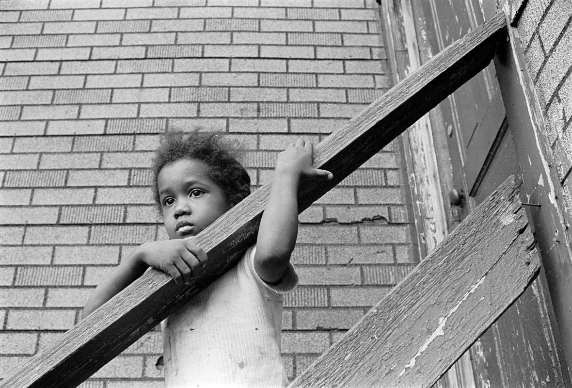 A child looks at the activity in the alley from the backstairs of the tenement where she lives.