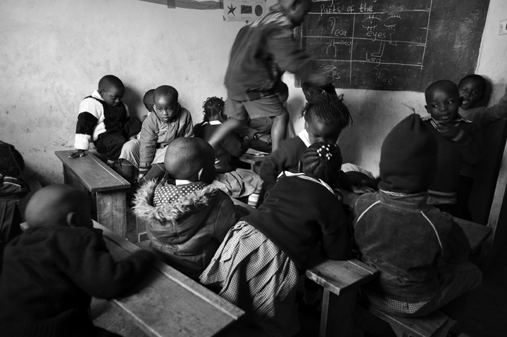 Nursery school students play while their teacher, at the front of the room, administers a test to three students at a time. {quote}I can't test the entire class at once because they would just play with and tear or stain the paperwork,{quote} says Cecilia Muringi.