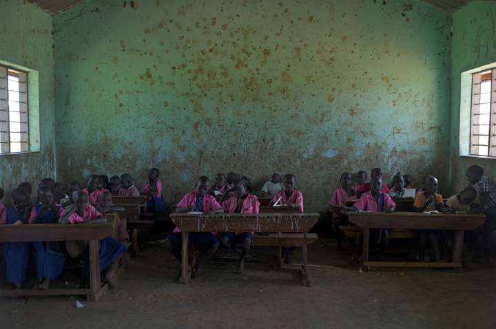 Primary one (P1) students at Acutomer Primary School wait for their teacher to return to the classroom; P1 students range in ages between six and eight. The room's windows have no glass so when it rains the students must move their desks to the center of the room. 