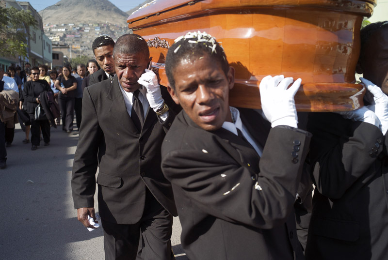 Eduardo Castillo answers his cellphone as his brother Victor Castillo struggles under the weight of a heavy coffin. Afro-Peruvian men (a vocational identity known as Camalenque) are highly sought to work as pallbearers - clad in tuxedos and white gloves at the most upscale funerals - under the belief that their skin-color lends an aura of elegance to the job. On May 18, 2010 Peru's Ministry of Culture denounced the practice of the Camalenque as racist and requested - to no avail - that the mortuary business end the service.
