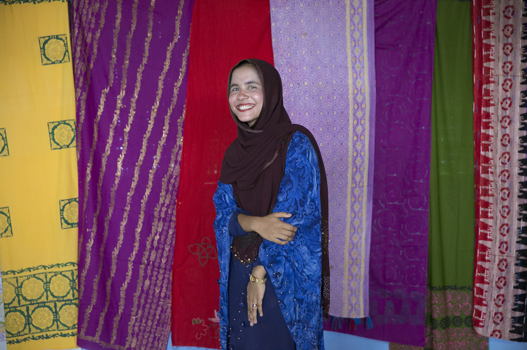 Lucky poses for a photo in front of a collection of Khanta blankets made by Rohingya women at the Women Friendly Center.  Lucky, 19, is taking a BRAC course on peace-building.  {quote}There are lots of groups working  here. If we work together we will be successful. If we are strong and raise out voices together we will united all the time. The biggest problem we face is domestic violence. They just think women are only good for cooking and raising families. If we are educated the situation will change.{quote}