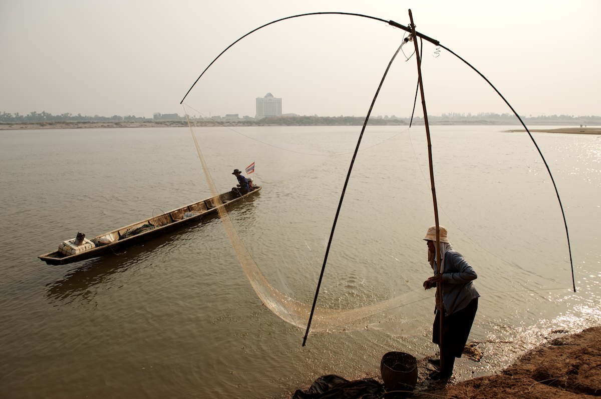 An elderly women net fishes on the bank of the Mekong River as her husband returns with nets he had set the previous night. 