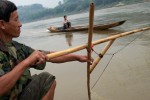 A fisherman uses a traditional net fishing technique to fish along the banks of the Mekong River. In the background a {quote}slow boat{quote} navigates locals and tourists upstream and downstream, stopping at tourist sites and villages. 