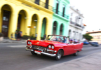Photo of young people driving a red car with colorful buildings in the background in Old Havana, Cuba. 