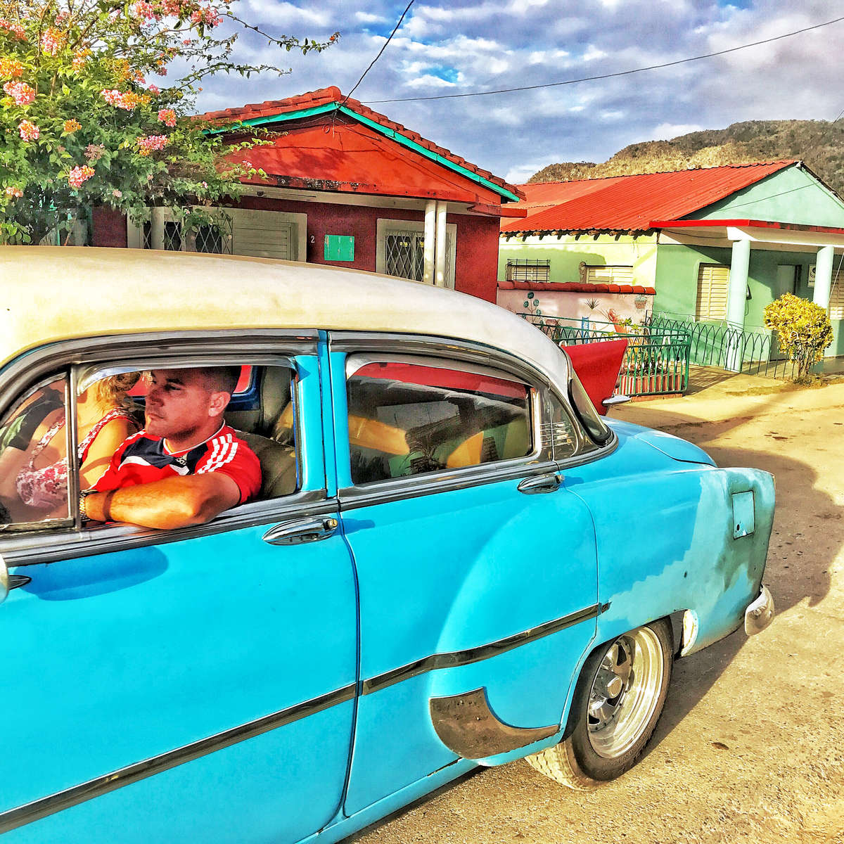 Photo of a Cuban couple driving their vintage 1952 Chevy in Viñales Valley, Cuba with colorful houses in the background.