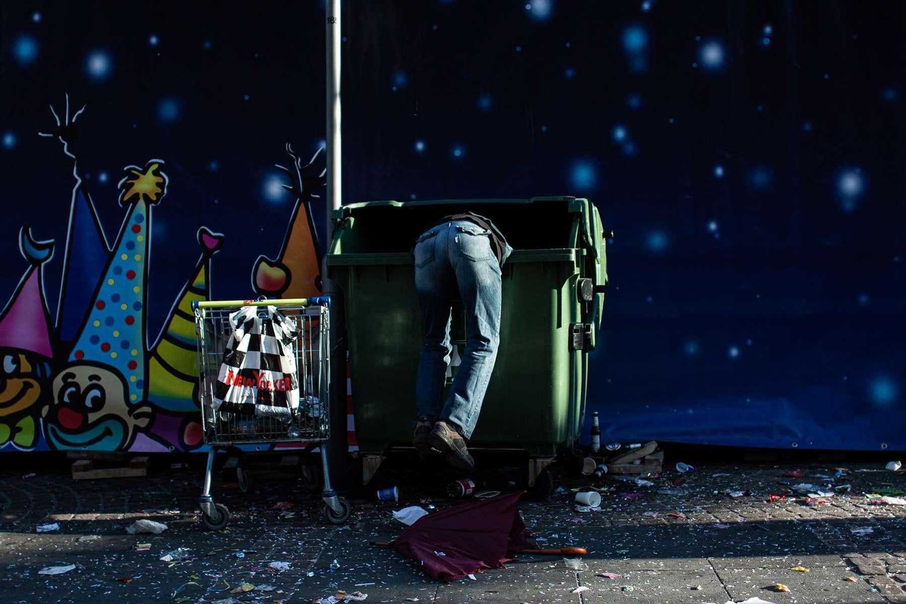 During the Rosenmontag carnival parade in Mainz in Germany a man is hanging and scavenging inside a trash can. His legs and the trash can are faintly lit by the evening sun
