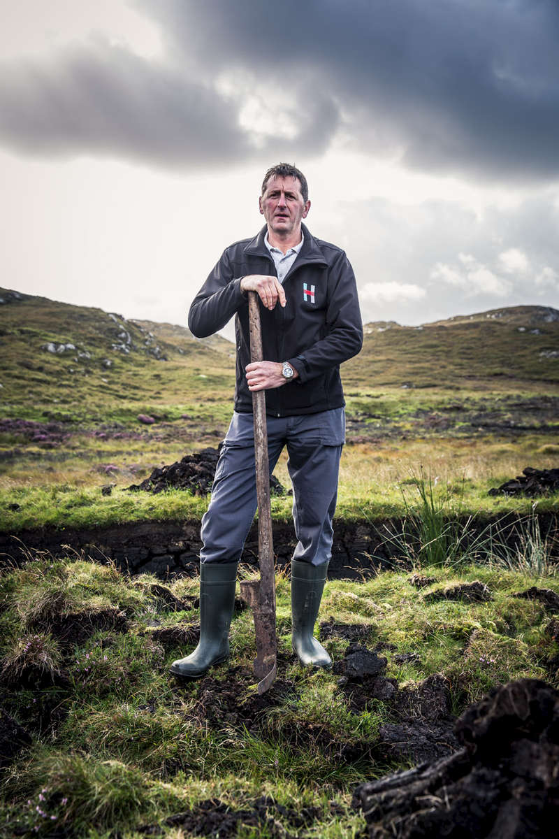 Distiller Stevie Passmore holding a tairsgeir peat cutter on a peat bank on a stormy day in the outer hebrides. 