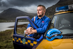 a local coastguard stands by his truck in the outer hebrides looking out to sea with mountains behind