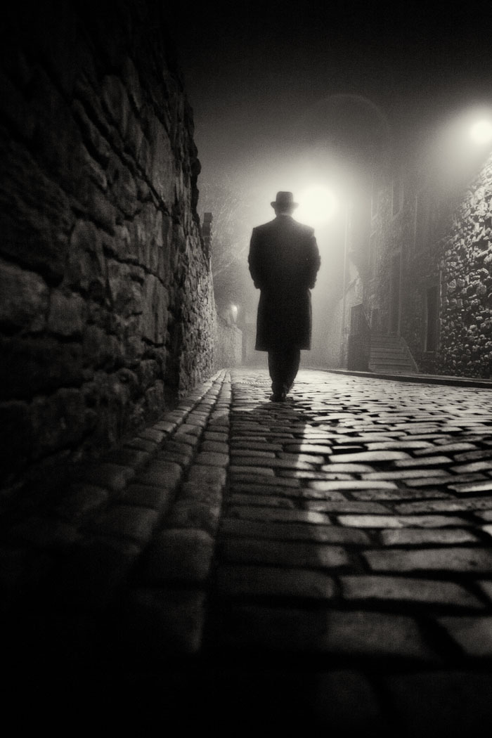 black and white. A man in a hat and coat walks down a cobbled street on a foggy night.