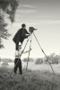a man stands on the shoulders of another to film with a vintage film camera resting on a home made wooden tripod