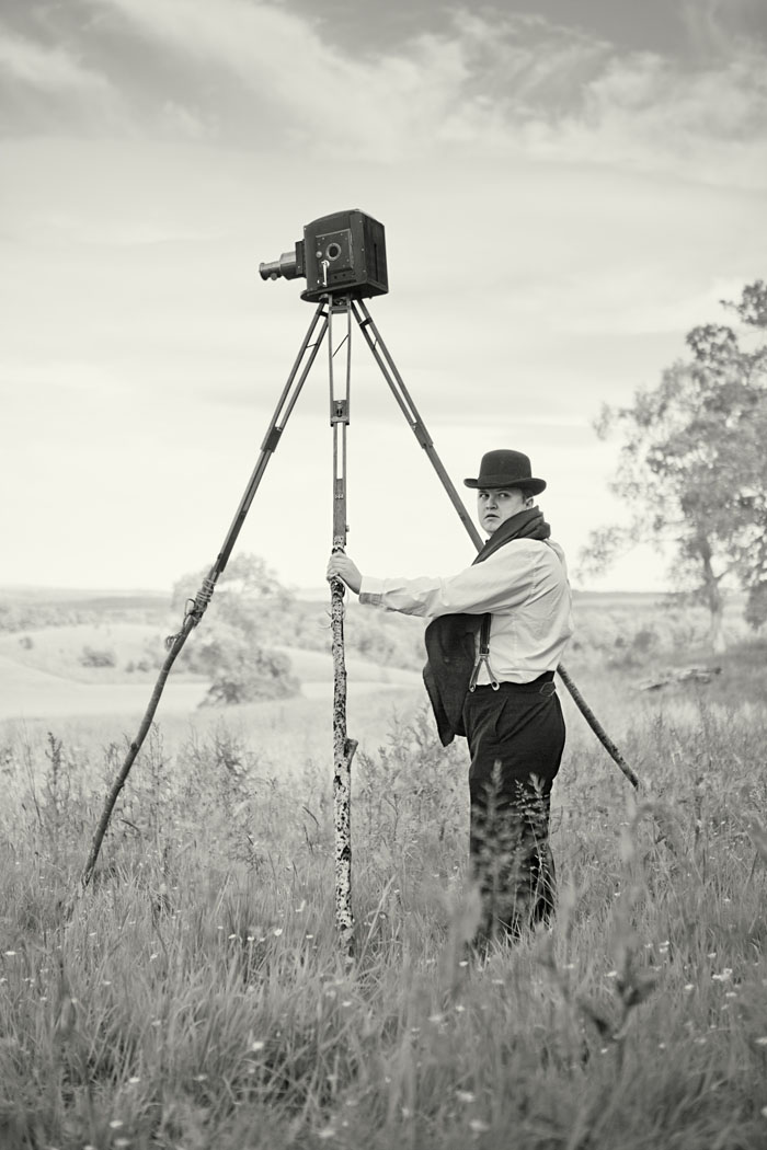 a man stands in a field holding a giant tripod with a film camera on it. Black and white