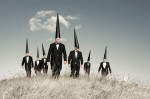 seven Conemen stride across a grassy landscape with black suits and black pointed hats