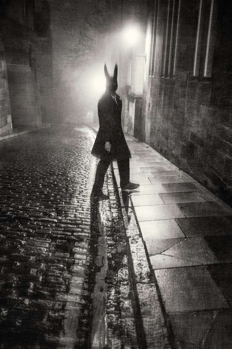 a man in a hare mask on a wet cobbled street at night