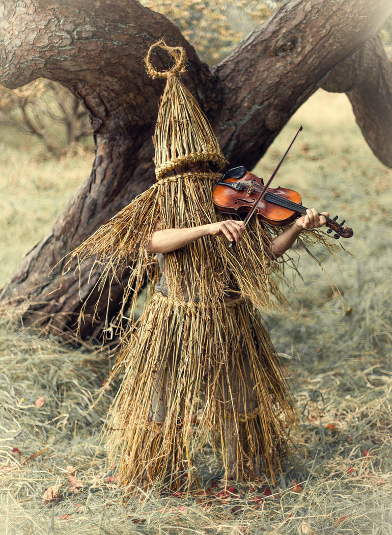 a skekler playing the fiddle in a forest. woman in a pagan witch outfit made of straw