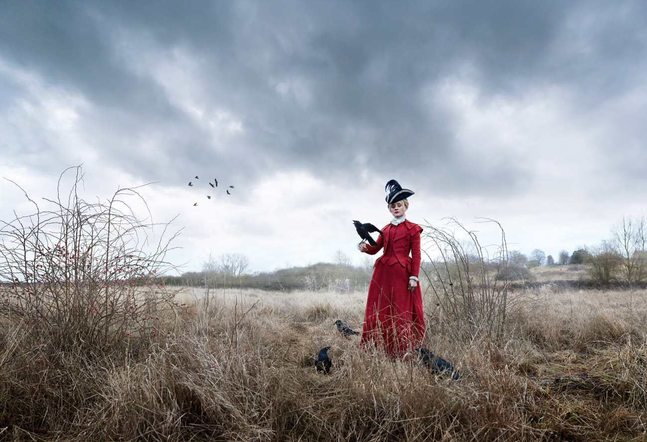 Anne Marie Duff. Woman in 17th century red dress standing in a thorny field with a crow on her arm