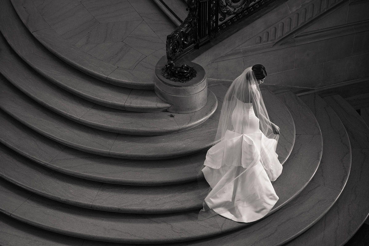 A bride stands on the grand staircase of San Francisco City Hall, a historic building with marble columns and a dome-shaped ceiling. The bride is wearing a white wedding dress and holding a bouquet of flowers, while sunlight streams through the windows and illuminates the scene. She is standing on the middle of the staircase, with one hand resting on the railing, and is looking towards the camera with a serene expression. The intricate details of the architecture of San Francisco City Hall are visible in the background, including the ornate patterns on the walls and the elegant chandeliers hanging from the ceiling. The image captures the elegance and beauty of a San Francisco City Hall wedding.