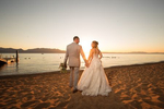 A bride and groom walk hand in hand along the shoreline of a sandy beach at Edgewood in Lake Tahoe as the sun sets behind them. The bride is wearing a flowing white wedding dress and holding a bouquet of flowers, while the groom is in a suit and tie. The calm waters of Lake Tahoe are visible in the background, with the mountains and trees surrounding the lake visible in the distance. The warm, golden light of the sunset illuminates the scene, casting a soft glow on the couple and the surrounding scenery. The peaceful and romantic setting of Edgewood provides the perfect backdrop for their wedding.