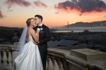 Bride and groom embracing during a sunset at Flood Mansion in San Francisco. The couple stands in front of the historic mansion, surrounded by the soft, warm colors of the setting sun. The iconic San Francisco skyline serves as a backdrop, creating a picturesque and romantic scene.