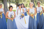 A group of bridesmaids stands together, laughing and having fun at a wedding at Nestldown, a scenic outdoor venue in the Santa Cruz Mountains. The bridesmaids are wearing matching light-colored dresses and holding bouquets of flowers. The bride is not in the picture, but her happy friends and family can be seen enjoying the moment together. The background is lush and green, with towering trees and foliage surrounding the group. The joyful and playful atmosphere of the moment is perfectly captured in this image.