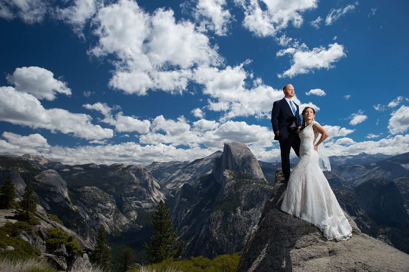 The happy couple is captured in a stunning image at Glacier Point observation point in Yosemite National Park. The bride is wearing a beautiful white wedding dress with a long train, and the groom is dressed in a stylish gray suit. They are standing at the edge of a cliff, with a breathtaking view of the Yosemite Valley and Half Dome in the background. The sun is setting, casting a warm glow on the couple and the surrounding landscape. A few wispy clouds are visible in the sky, adding to the natural beauty of the scene. This image captures the beauty and romance of a wedding held in the midst of one of America's most iconic national parks.
