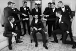 A group of groomsmen are gathered in a room, getting ready for a wedding at Lafayette Park in San Francisco. The groom is standing in the center of the group, wearing a black suit and a white shirt, and holding a boutonniere. The groomsmen are also wearing black suits with white shirts and black ties. They are all focused on getting ready, with some tying their ties and others buttoning their jackets. The room is well-lit with natural light, and a window in the background shows a view of the park outside. The groom and his groomsmen are excited and ready to celebrate the wedding day together.