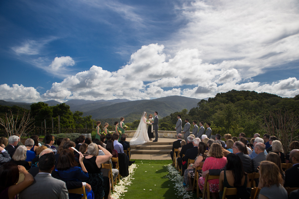 A couple stands at the front of a lush green lawn at Holman Ranch in Carmel, California, surrounded by their family and friends who are seated on chairs arranged in rows.  A white wooden arch with flowers and greenery is visible behind them, and the mountains and trees of Carmel Valley are visible in the distance. The sun is shining, casting a warm and inviting light on the scene. The serene and picturesque setting of Holman Ranch provides a beautiful backdrop for their wedding ceremony.