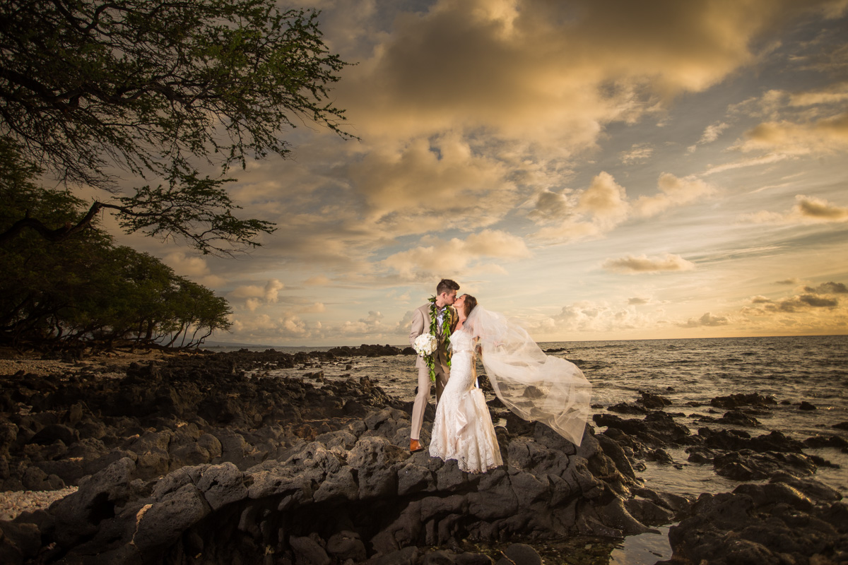 A stunning couple is captured in a sunset portrait at Mauna Kea Resort, a luxurious beachfront hotel in Hawaii. The bride is wearing a beautiful white wedding gown with delicate lace details, while the groom is dressed in a sharp suit with a boutonniere on his lapel. The image is taken on a sandy beach, with palm trees and a clear blue sky in the background. The sun is setting over the ocean, casting a warm orange glow across the scene. The couple is holding hands and gazing lovingly into each other's eyes, with smiles on their faces. This image captures the beauty and romance of a tropical destination wedding, as well as the stunning natural scenery of Hawaii.