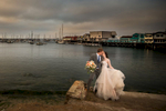 A bride and groom are seen in the historic downtown area of Monterey, California, posing for wedding photos at sunset. The bride is wearing a white wedding dress with a bouquet of flowers, while the groom is in a suit and tie. They are standing on a cobbled street with a row of colorful old buildings in the background. The sun is setting, casting a warm, golden light on the scene. The historic architecture of the buildings and the colorful street make for a picturesque backdrop for their wedding photos. The couple had their wedding at Perry House, a nearby historic venue known for its charming gardens and Victorian architecture.