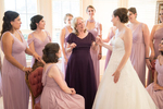 The mother of the bride is captured in a heartwarming moment with her daughter and bridesmaids at a wedding held at Kohl Mansion, a historic venue in Burlingame, California. The bride is dressed in a stunning white wedding gown with intricate lace details, while the bridesmaids are wearing elegant pale pink dresses. The mother of the bride is dressed in a stylish silver-gray gown and standing in the center of the group, with her arm around her daughter. They are all smiling and laughing, with a beautiful garden in the background. The image captures the love and bond between the mother and daughter, as well as the joy and camaraderie of the bridesmaids on this special day.