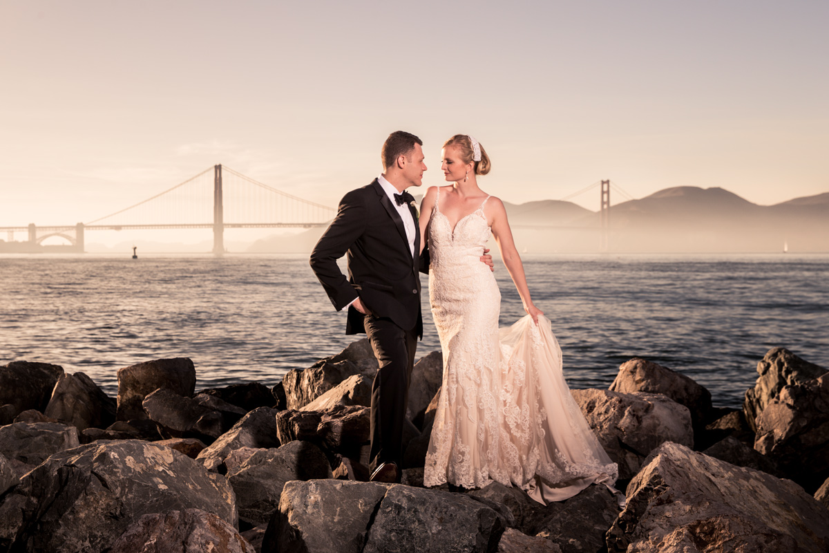 An outdoor portrait captures the silhouette of a newlywed couple against a stunning sunset backdrop. The groom, dressed in a navy suit, holds the bride's hand as she wears a flowing white wedding gown. The scene takes place at the St. Francis Yacht Club, where the couple's wedding ceremony took place.