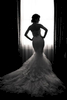  A black and white photo of a bride standing in front of a large window with a silhouette effect. The bride's gown and veil are highlighted against the bright window light. The background is blurred out, creating a dreamy and romantic feel.