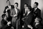 A group of groomsmen are gathered in a luxurious hotel room at the Westin St. Francis hotel in San Francisco, California. The room is elegantly decorated with plush furnishings, and a large window in the background offers views of the city skyline. The groomsmen are dressed in sharp black suits with white shirts and black ties, and are in the process of getting ready for the wedding. Some are adjusting their ties, while others are putting on jackets or shining their shoes. The image captures the excitement and anticipation of the groomsmen as they prepare to celebrate the wedding day with their close friend.
