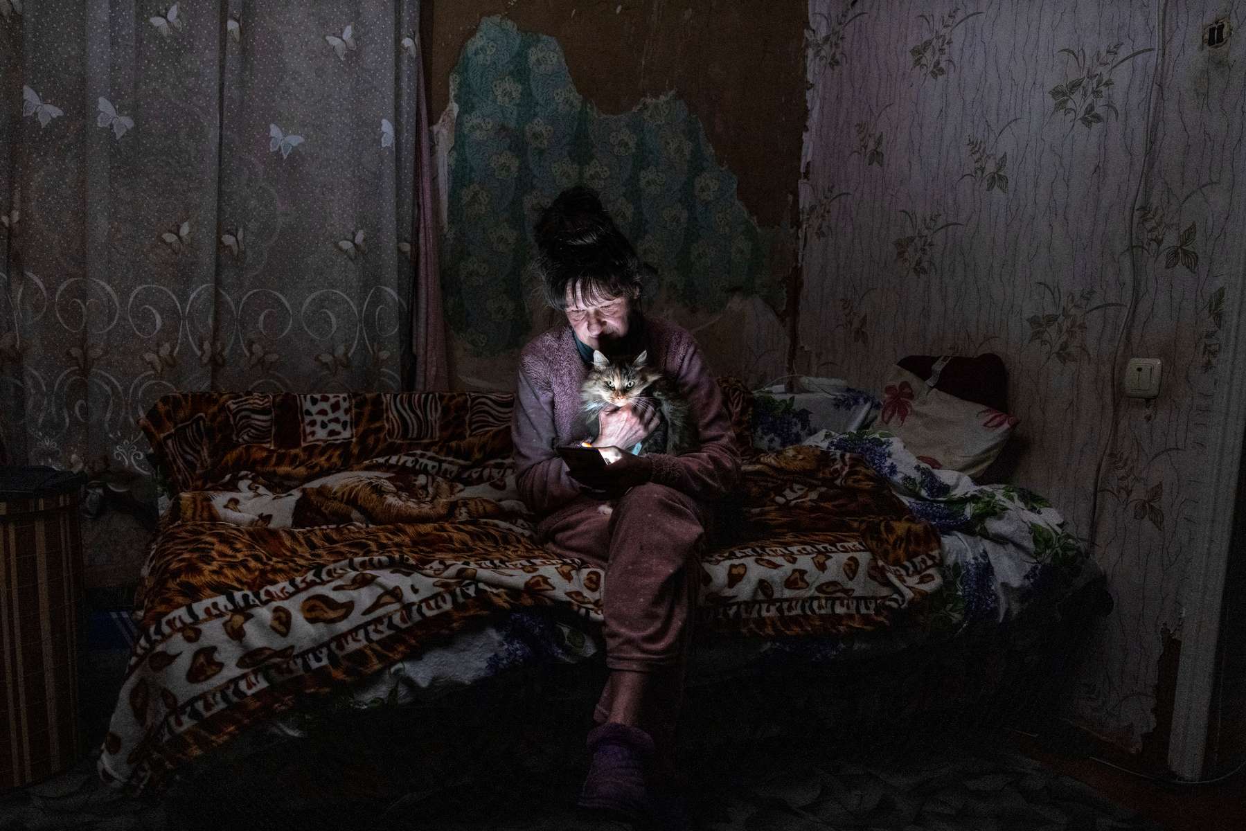BORODYANKA, UKRAINE - Tetyana Safonova, 61, sits with her cat Asya as she looks at her mobile phone during a power outage on October 20, 2022 in Borodyanka, Ukraine. Tetyana wasn’t able to buy candles in town because of the sudden demand given the unpredictable power cuts that the government has imposed around Ukraine. Restricted power supplies and limited electricity started today so that energy companies could repair power facilities hit by a wave of recent Russian air strikes. (Photo by Paula Bronstein /Getty Images)