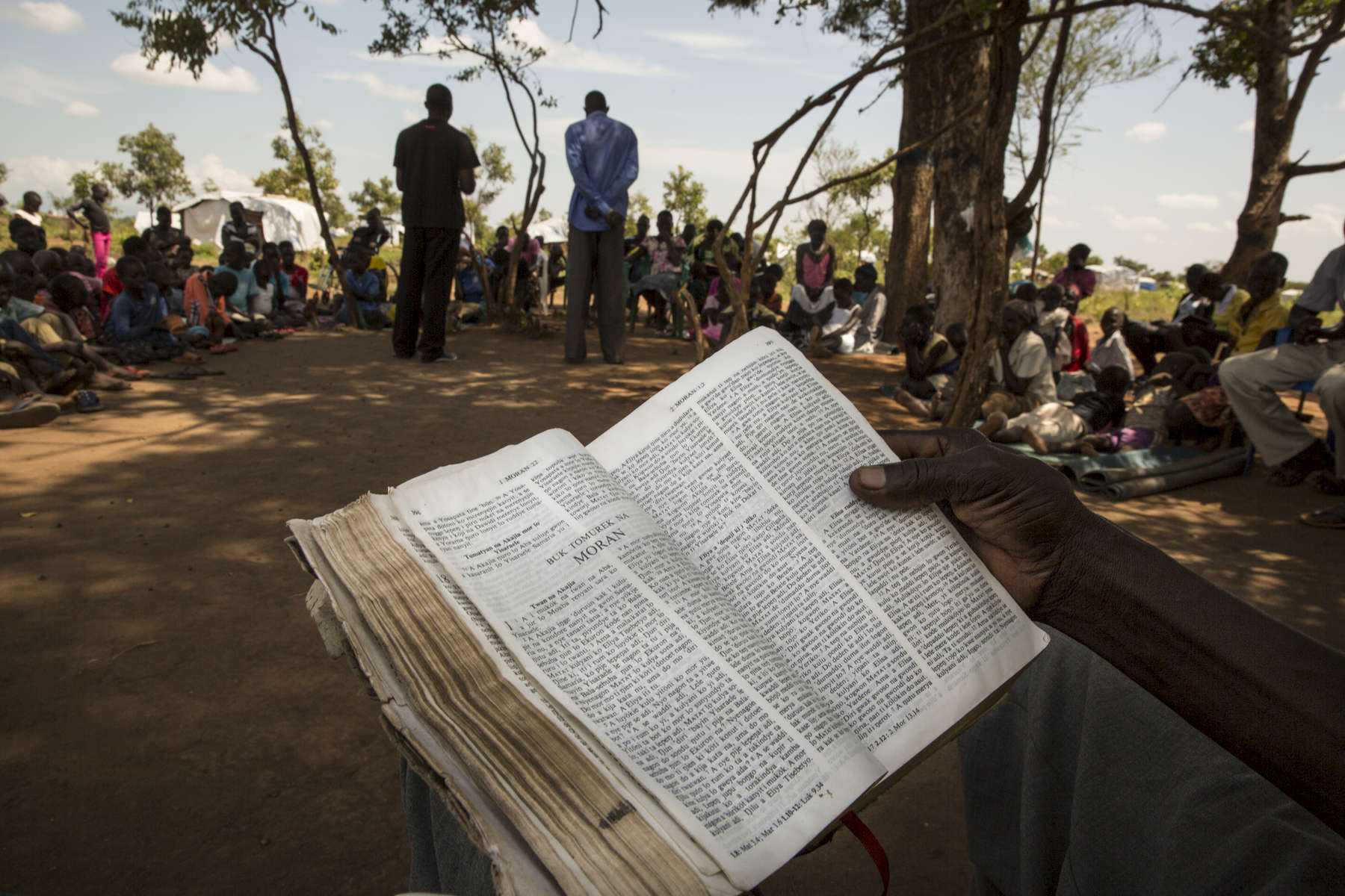 ADJUMANI DISTRICT-UGANDA:  During a Catholic Sunday prayer service held under the shade of a tree, refugees sit and pray as priests deliver their sermon.The Onward Struggle:  A refugee crisis in Uganda deepens as South Sudanese Refugees are forced to leave their country behind. The outbreak of violence in the capital Juba last July created a humanitarian crisis in northern Uganda as thousands of South Sudanese sought refugee there. The country is hosting the lion’s share of South Sudanese refugees, with 373,626, more than a third of them arriving since early July.The fighting was a major setback to peace efforts in South Sudan, coming as the troubled new nation prepared to celebrate its fifth anniversary, amid a short lived peace deal between supporters of President Salva Kiir and former First Vice President Riek Machar. South Sudan now joins Syria, Afghanistan and Somalia as countries which have produced more than a million refugees. While some South Sudanese may attempt to head for Europe, the numbers within east Africa are comparable in scale to recent refugee flows to Europe from the Middle East, and their traumatic experiences due to war are often just as hellish. More than 85 percent of the refugees in this recent influx are women and children. Many children have lost one or both of their parents, some forced to become primary caregivers to siblings.With the large influx of refugees in July 2015, relief agencies had to implement stringent food rationing in the refugee settlements. Currently the international humanitarian organizations lack the necessary funds to meet the needs of the more than 200,000 refugees.